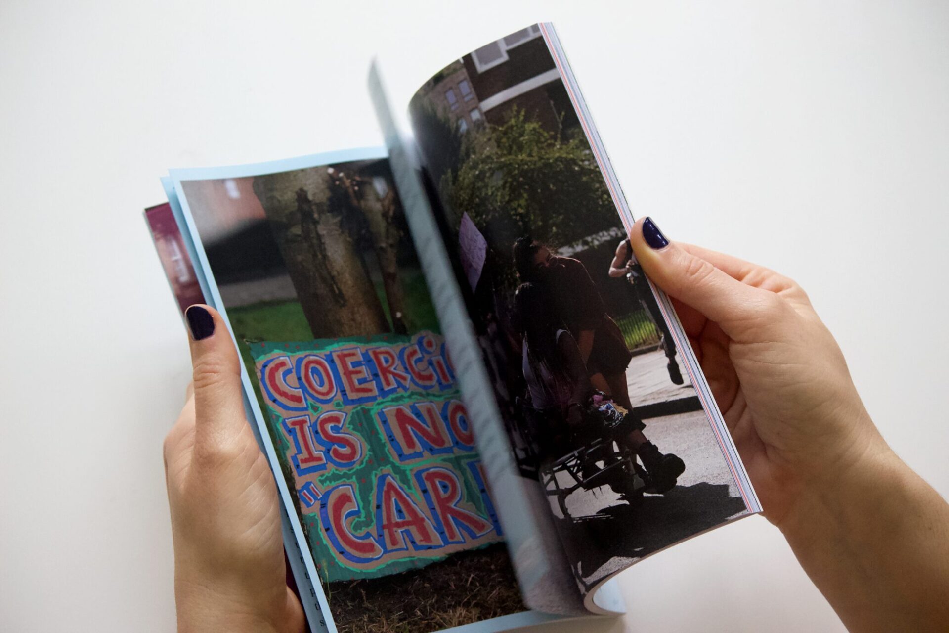 The photo shows hands holding the zine, an inner page displayed. The pay is a photograph of a protest with the slogan 'coercion is not "care"'